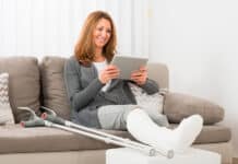 nainen istuu kipsi jalassa sohvalla kädessään tablettitietokone